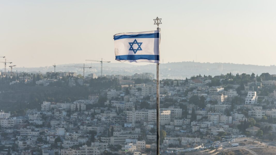 israeli national flag waving on the top of mount of olive with background of residential houses in jerusalem, israel